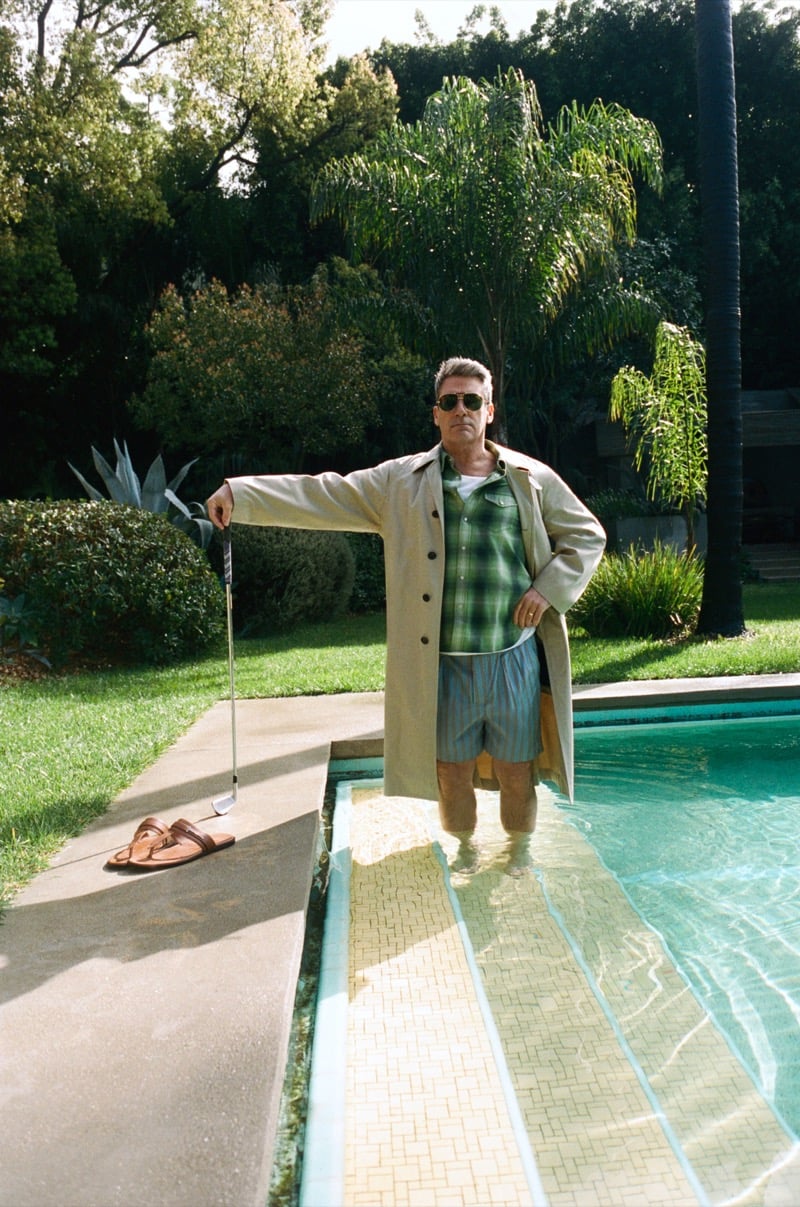 Jon stands poolside wearing aviator sunglasses, a belted trench coat, checked shirt, and striped shorts