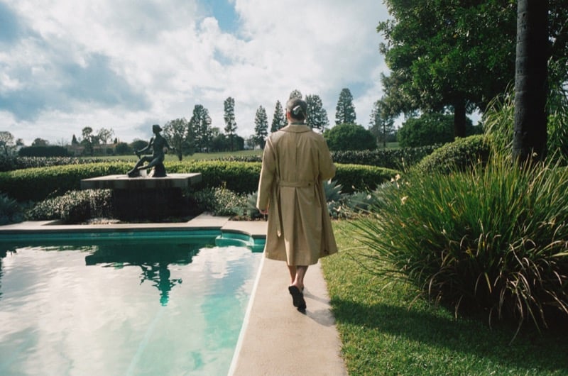 Jon walks along a pool in a belted trench coat