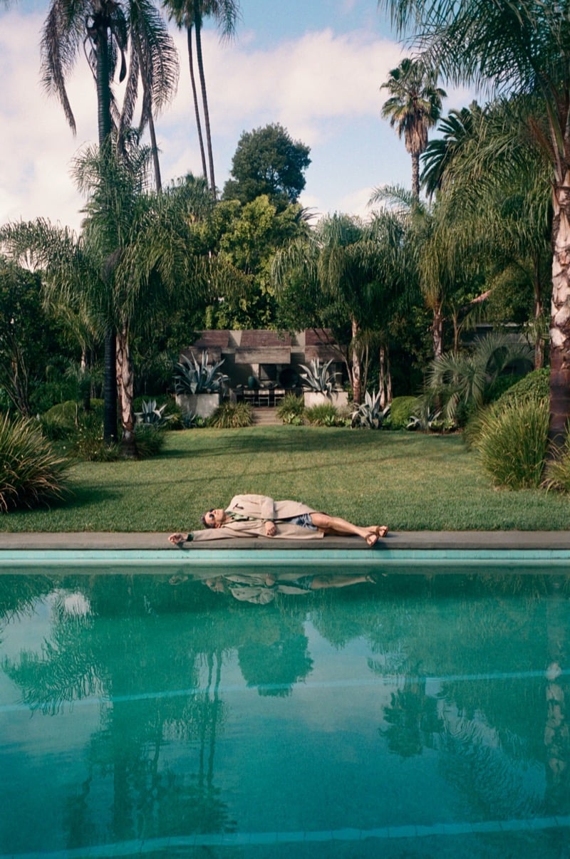 Jon lies poolside in a trench coat and striped shorts