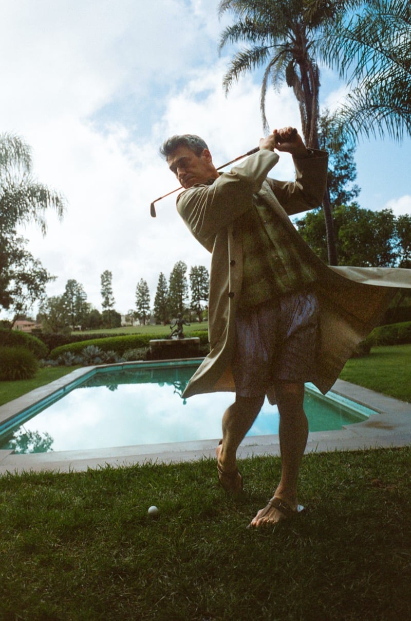Jon swings a golf club poolside wearing a trench coat and striped shorts