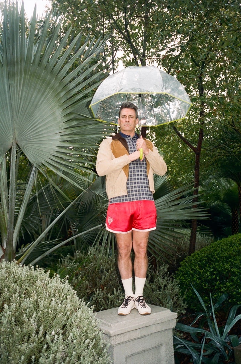 Jon stands on a garden plinth holding an umbrella wearing a suede jacket, checked polo shirt, and red satin shorts