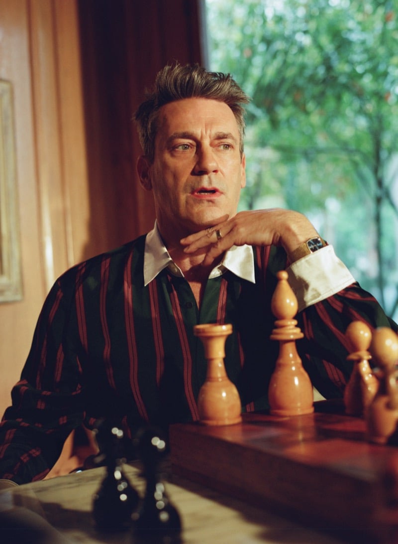 Jon rests his chin on his hand at the chess table in a striped silk shirt