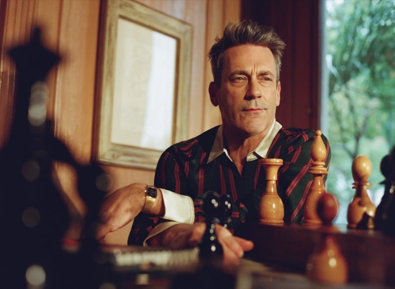 Jon leans back at a chess table holding a chess piece in a striped silk shirt