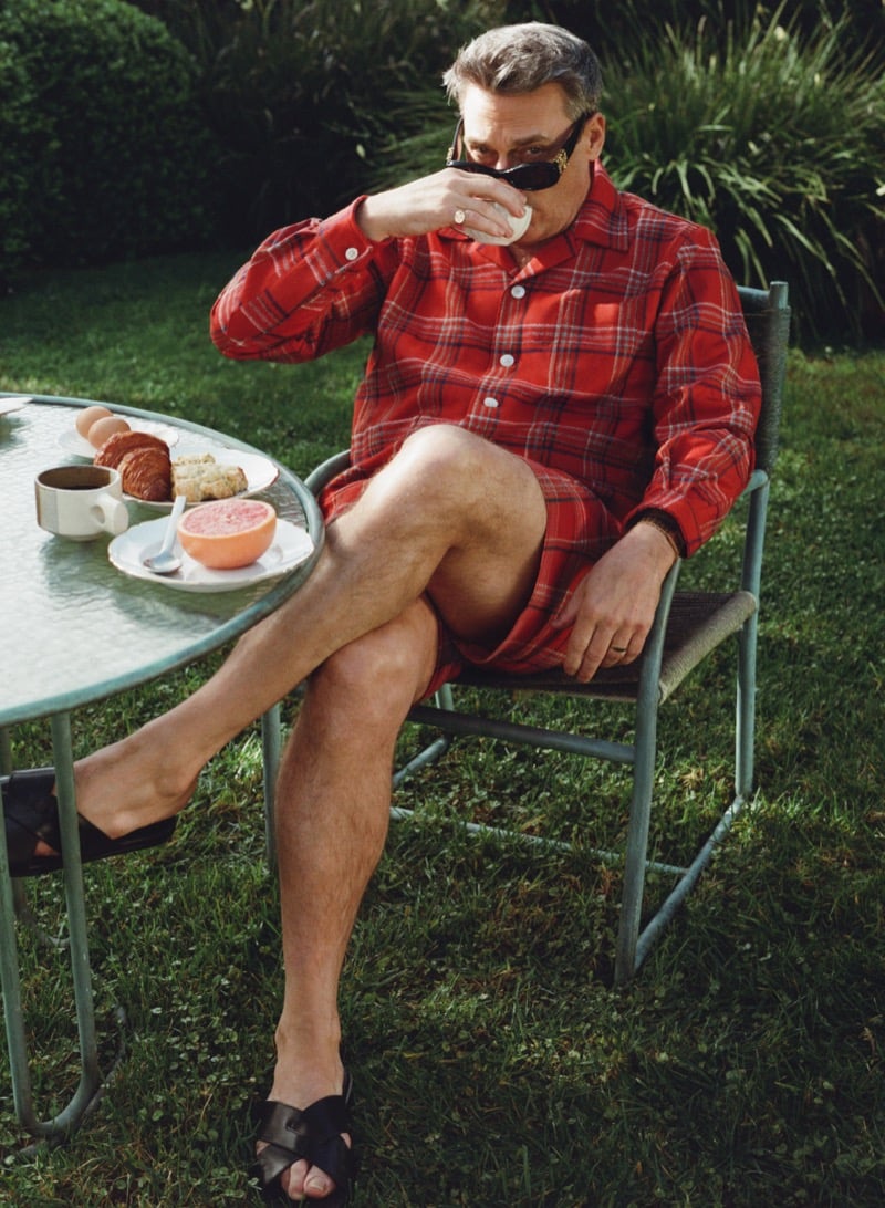 Jon sits in a garden chair drinking from a glass wearing a checked wool overshirt, shorts, and leather slides