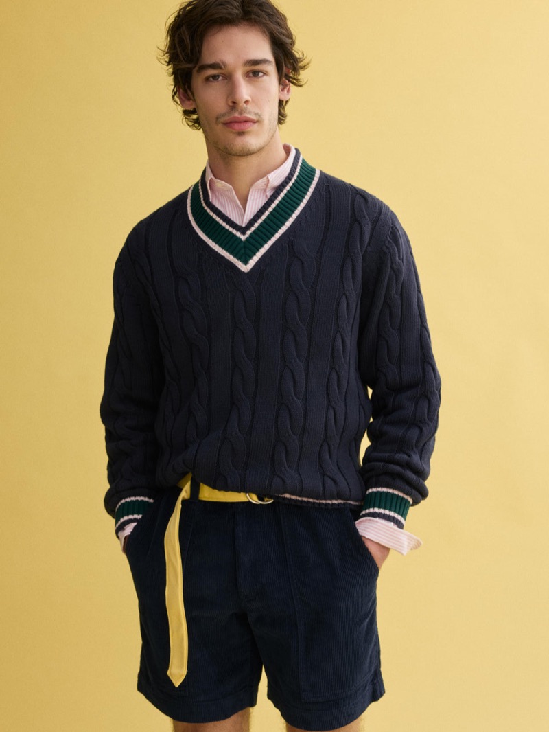 Klevi wears a J.Press x Alex Mill navy cable-knit cricket sweater with green tipping over a pink striped shirt, paired with navy corduroy shorts and a yellow D-ring belt