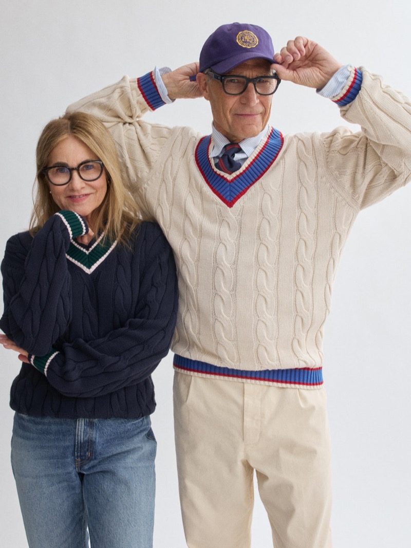 Michael wears a J.Press x Alex Mill cream cable-knit cricket sweater with red and blue tipping, a striped shirt, knit tie, and a purple J.Press baseball cap, beside a woman in a navy cricket sweater