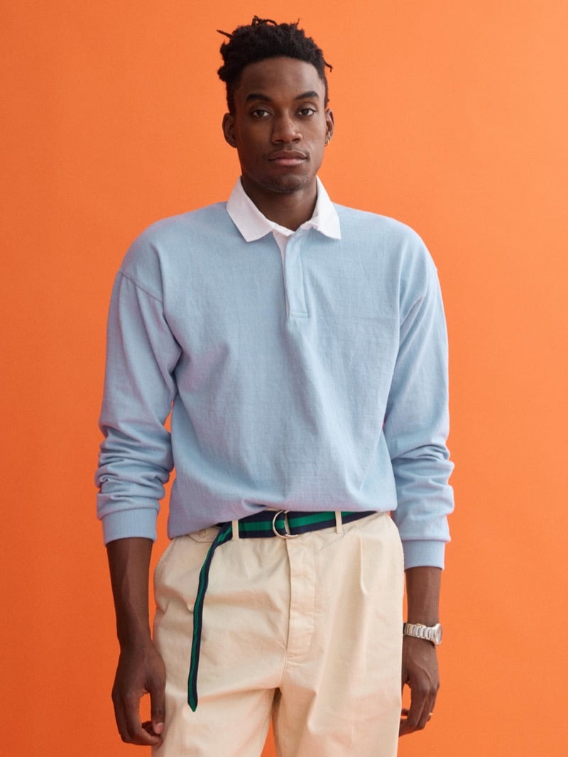 Herzen wears a J.Press x Alex Mill light blue rugby shirt with a white collar, paired with cream pleated chinos and a navy and green D-ring belt