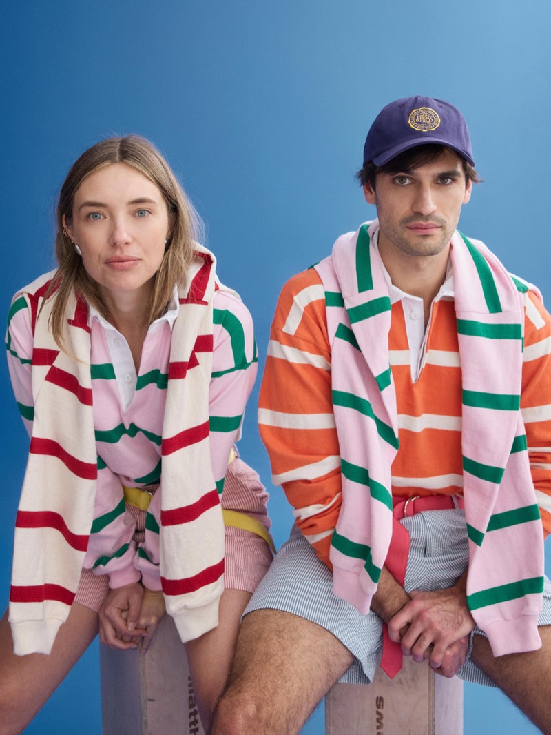 Klevi wears a J.Press x Alex Mill pink, orange, and green striped rugby shirt with striped seersucker shorts and a navy J.Press baseball cap, seated beside a female model in a matching pink, red, and green striped rugby