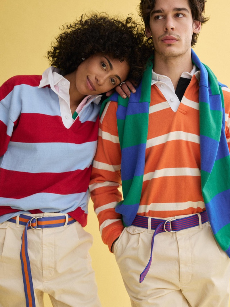 Klevi wears a J.Press x Alex Mill orange, green, and blue striped rugby shirt with cream pleated chinos and a purple D-ring belt, beside a female model in a red and light blue striped rugby