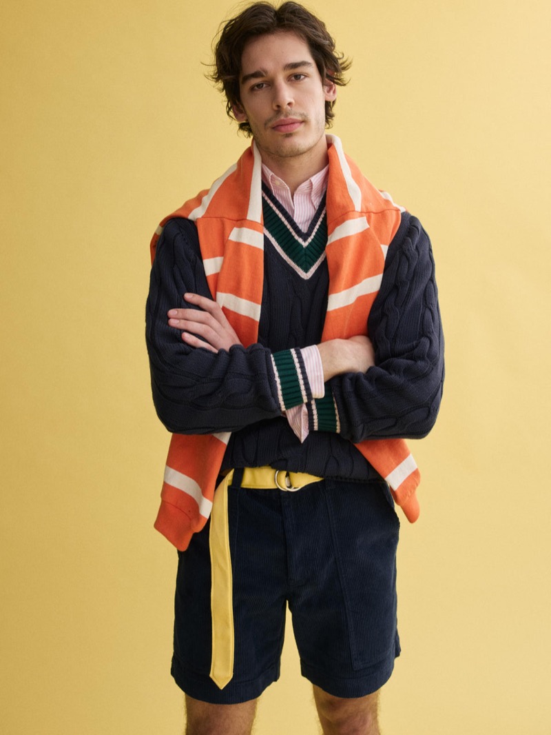 Klevi wears a navy J.Press x Alex Mill cable-knit cricket sweater with an orange and cream striped rugby draped over his shoulders, navy corduroy shorts, and a yellow D-ring belt