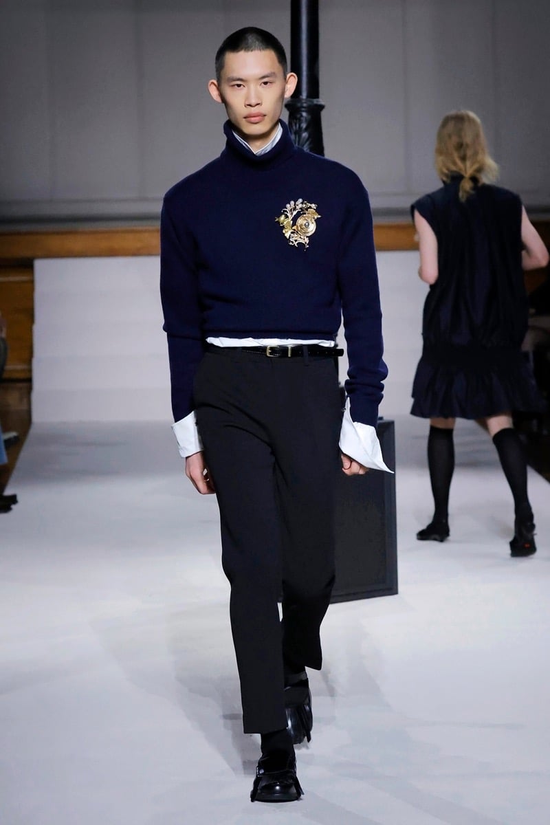 Kent & Curwen fall-winter 2026 navy rollneck sweater with gold crest brooch over a white dress shirt and black cropped trousers