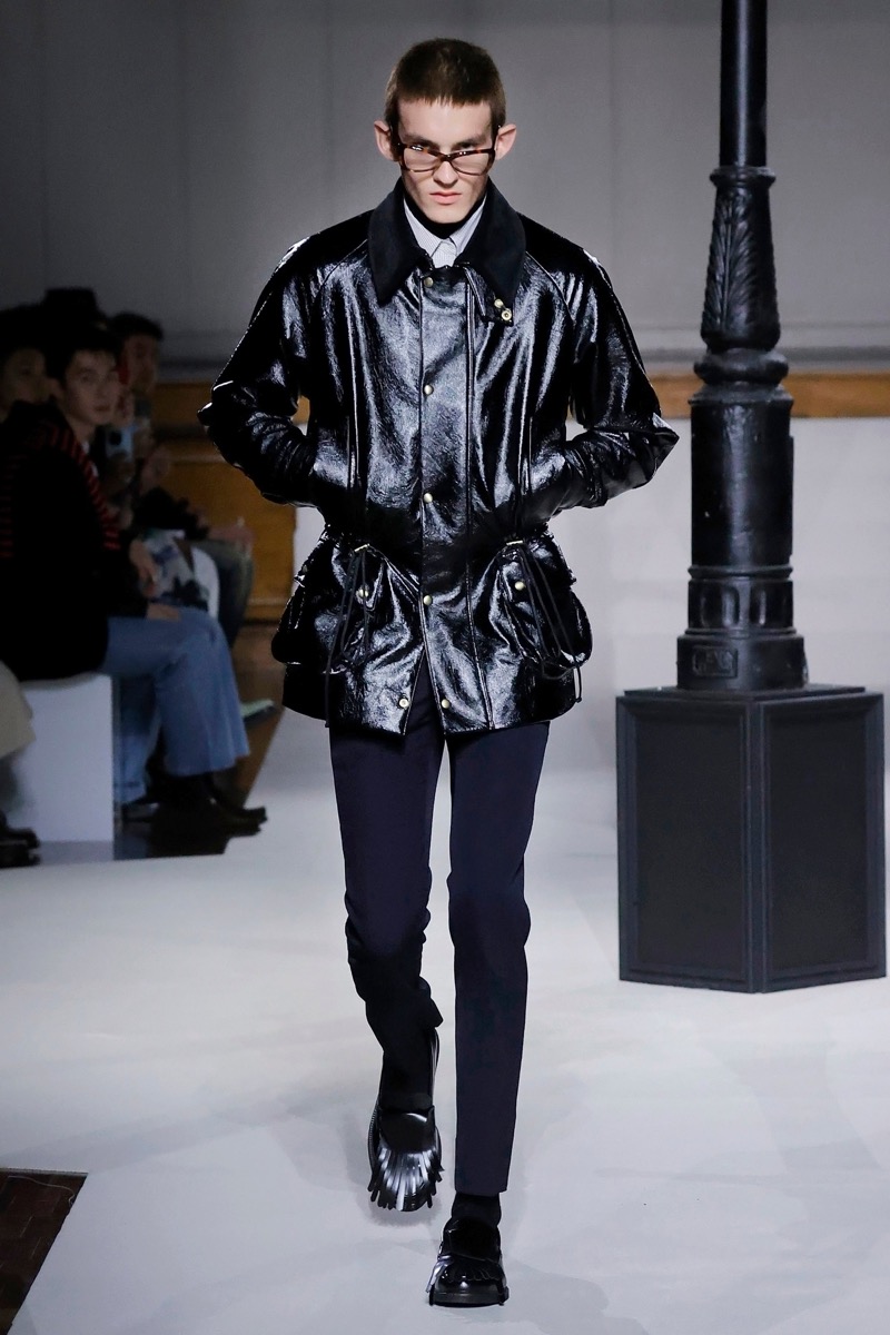 Kent & Curwen fall-winter 2026 black patent leather jacket with brass hardware over a white shirt and navy trousers