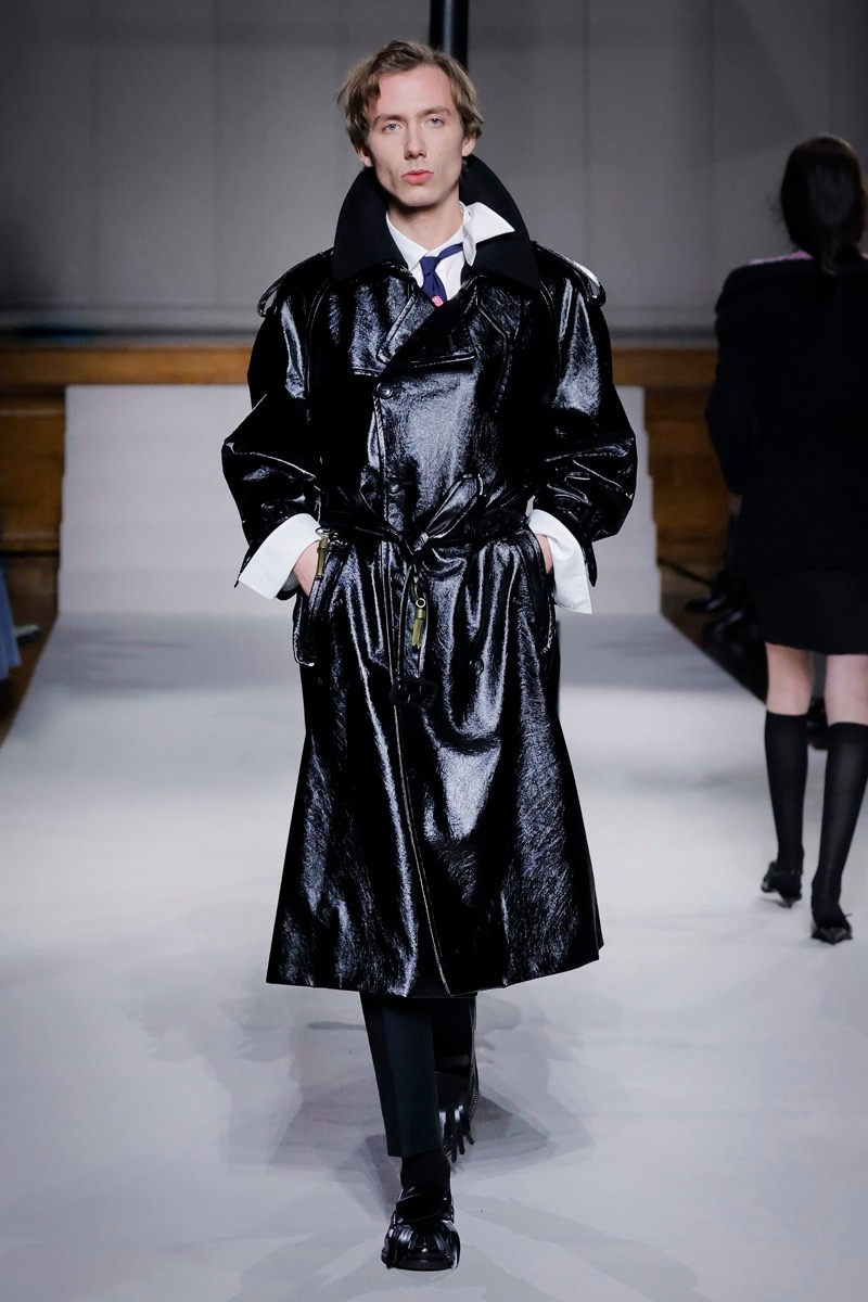 Kent & Curwen fall-winter 2026 black patent leather belted trench coat over a white shirt with a striped tie and charcoal trousers