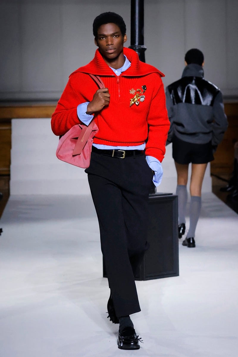 Kent & Curwen fall-winter 2026 cherry red half-zip sweater with gold embroidered brooch over a light blue shirt and black wide-leg trousers with a pink bag
