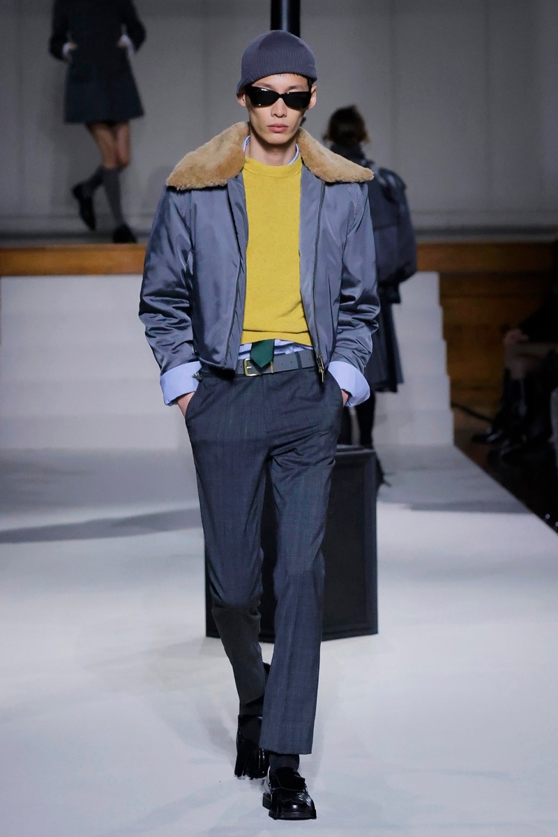 Kent & Curwen fall-winter 2026 gray bomber jacket with shearling collar over a yellow sweater and charcoal trousers with a navy beanie and sunglasses