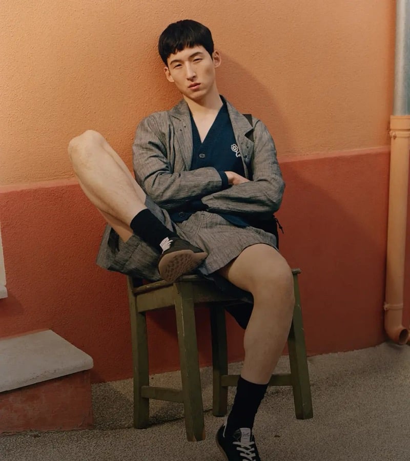 Woosang sits on a small green wooden stool against a peach-pink wall in a gray linen blazer layered over a dark navy cardigan, with shorts, black crew socks, and black sneakers for Kenzo's pre-fall 2026 campaign