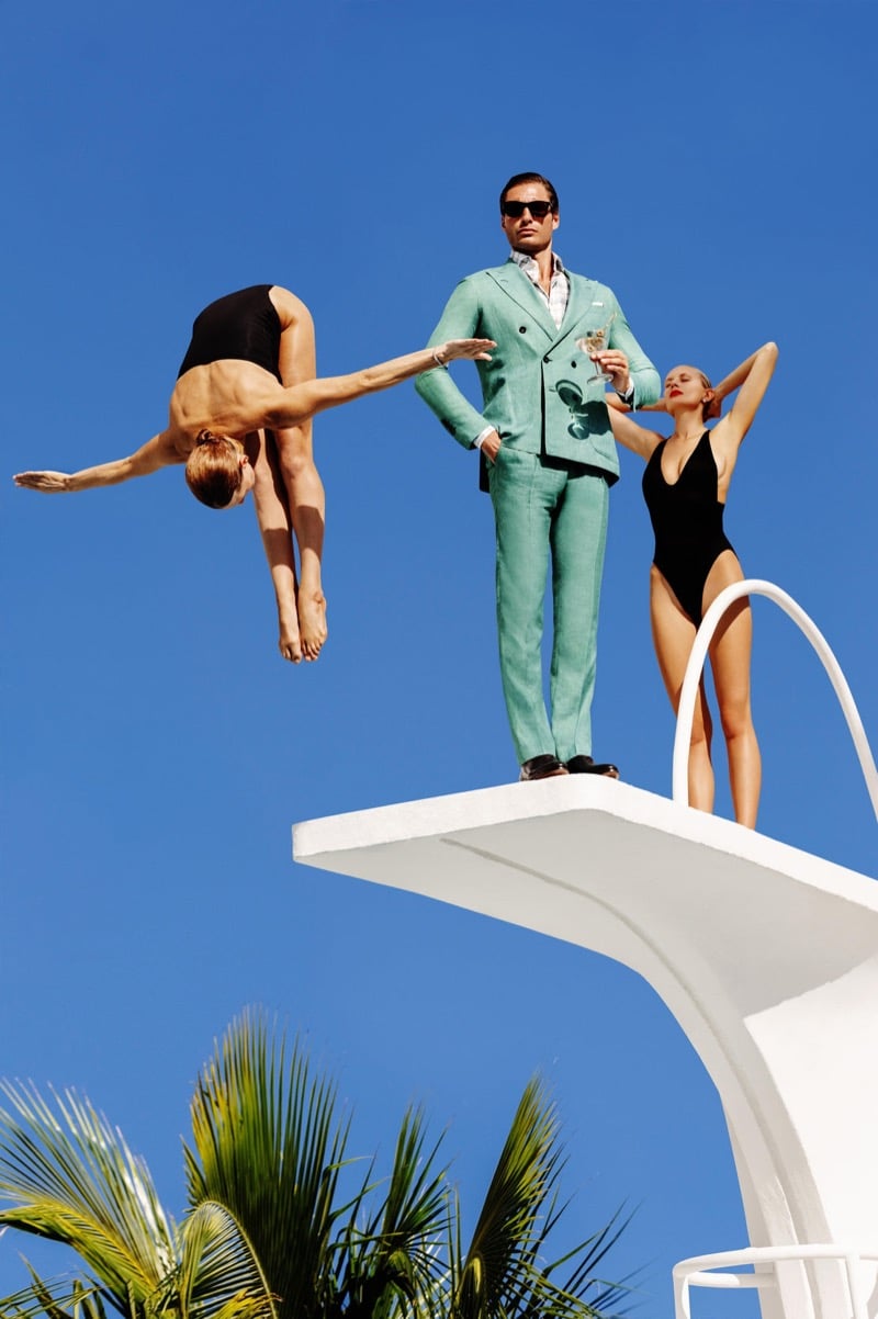 Christopher stands on a diving board in a mint-green double-breasted suit with a champagne glass as swimmers dive beside him