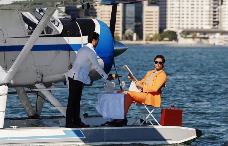 Christopher reclines on a seaplane pontoon in an orange suit reading a newspaper while a waiter serves champagne