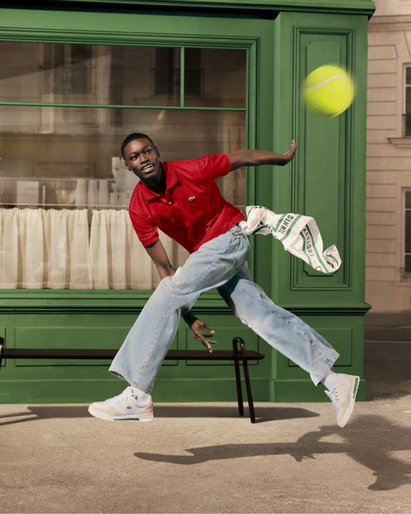 Diakhate lunges to return a tennis ball on a Paris street in a red Lacoste polo, light wash jeans, and white Lacoste sneakers, with a Lacoste tennis towel flying behind him for the "Life Is a Beautiful Sport" campaign