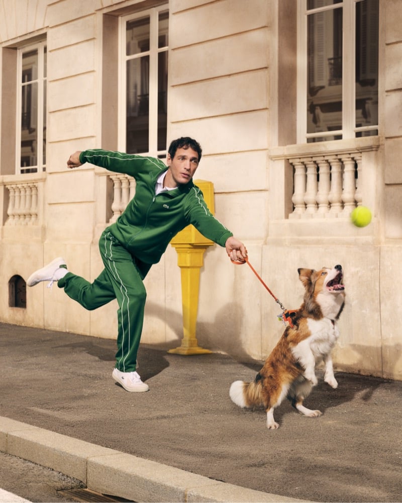 Talbourdet mid-stride on a Paris street in a green Lacoste tracksuit with white piping, holding a dog on an orange leash while a tennis ball flies overhead for the "Life Is a Beautiful Sport" campaign