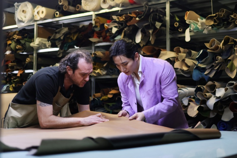 Jun-ho and a Berluti artisan examine a piece of leather at the cutting table surrounded by rolled hides