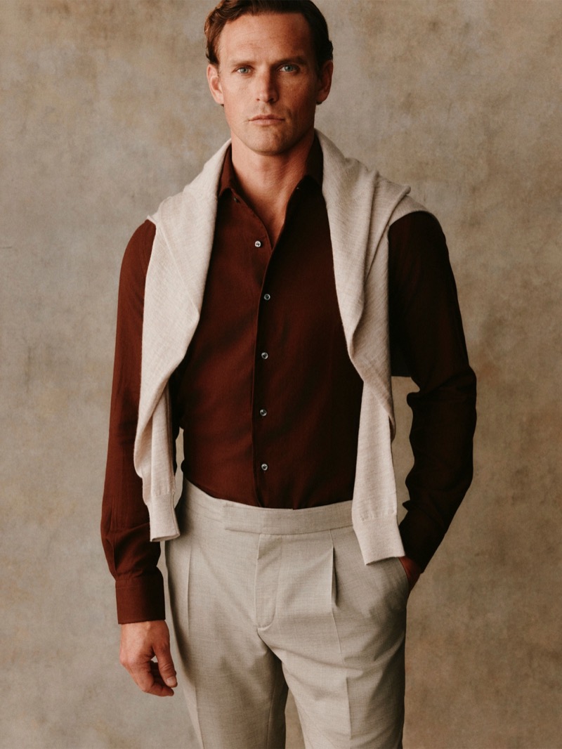 Guy wears a burgundy shirt with a stone sweater draped over his shoulders and stone trousers