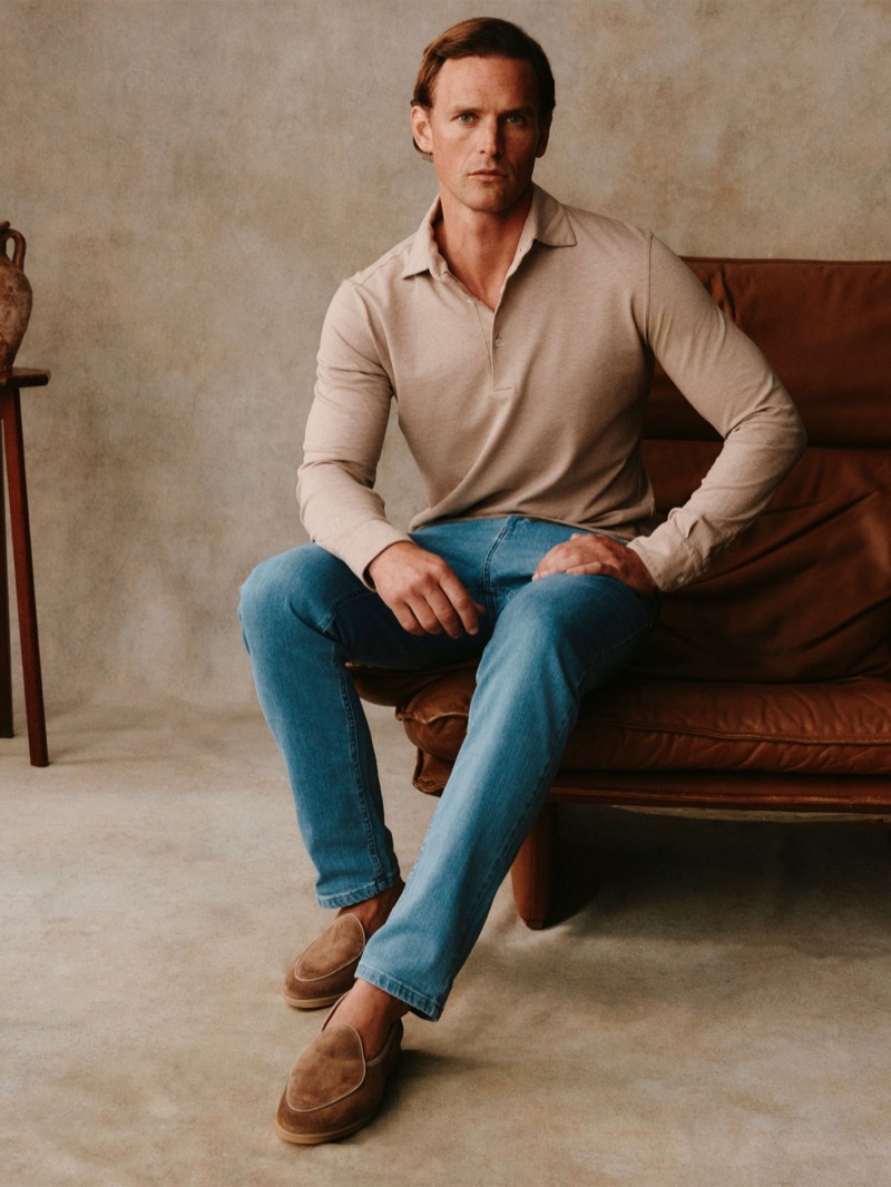Guy sits in a beige long-sleeve polo with blue jeans and suede loafers