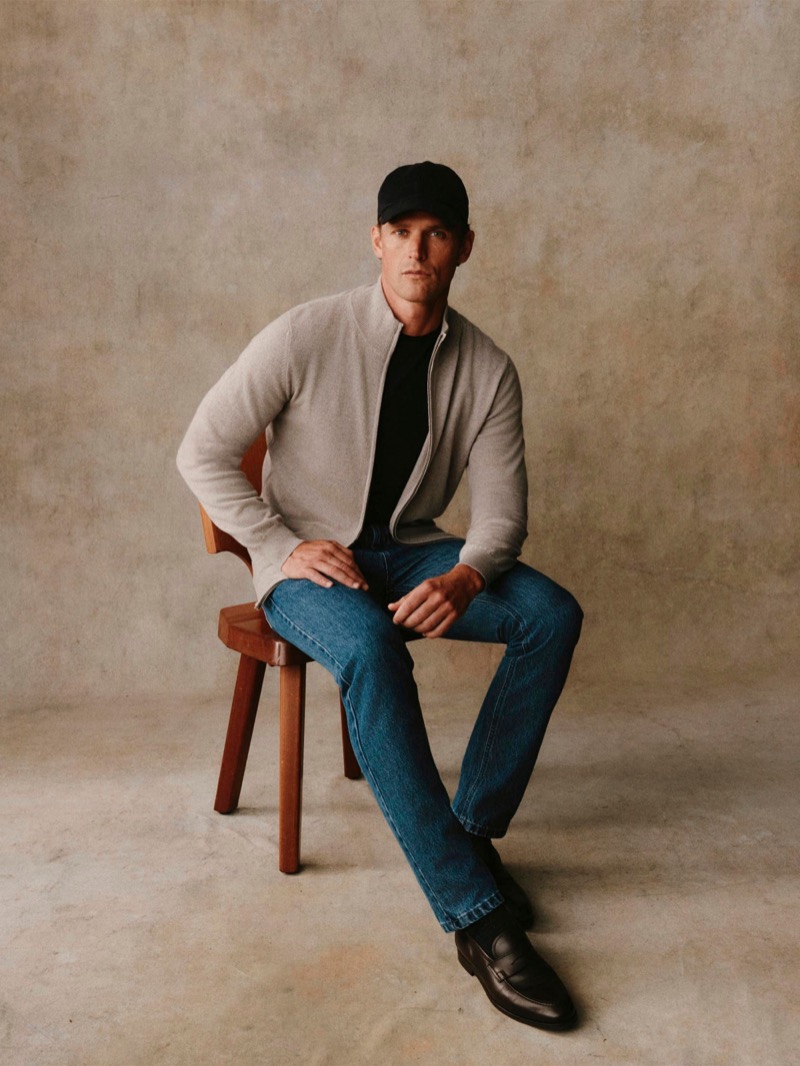 Guy sits in a stone zip-front cardigan over a black tee with jeans and a cap
