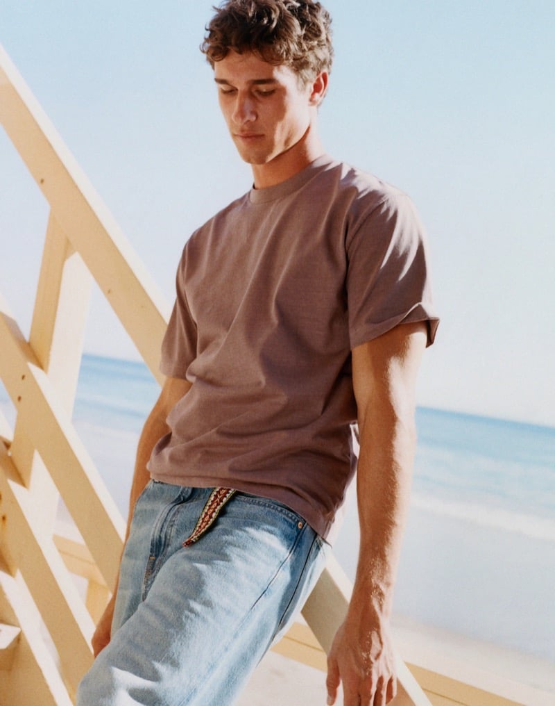 Kai wears a dusty mauve T-shirt and light-wash jeans with a woven braided belt beside a wooden beach structure