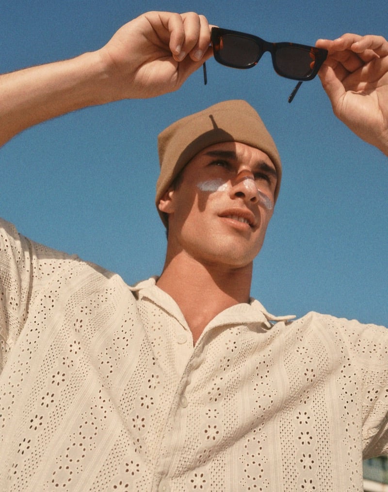 Kai holds black sunglasses overhead wearing an eyelet-embroidered cream camp shirt and tan fisherman beanie with sunscreen on his nose