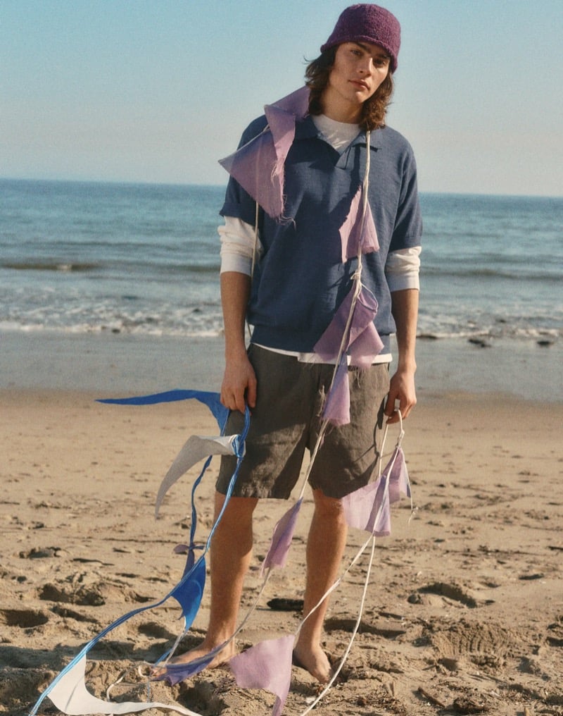 Sava stands barefoot on the beach in a navy johnny-collar polo over a white long-sleeve shirt with dark olive pull-on shorts and a purple bucket hat, holding colorful kite streamers