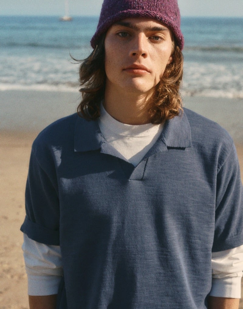 Sava looks directly at the camera on the beach in a navy slub-knit Johnny collar polo layered over a white long-sleeve shirt and a purple knit beanie
