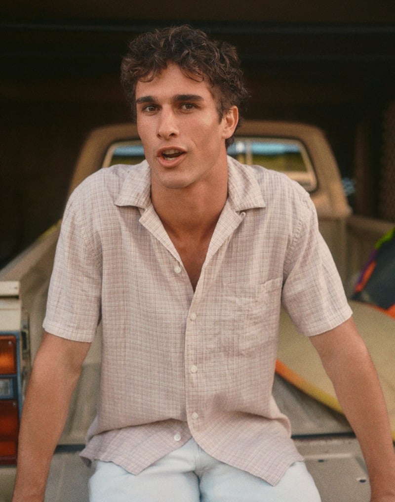 Kai sits on the bed of a vintage pickup truck in a grid-check camp shirt with the collar open