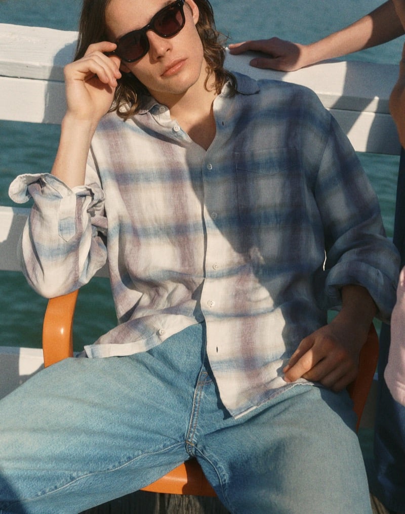 Sava sits on a boat deck wearing a blue ombre plaid linen shirt and black sunglasses with light-wash jeans