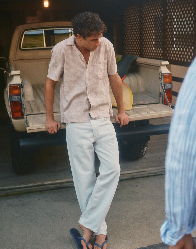 Kai leans against a pickup truck tailgate in a grid-check camp shirt with light-wash jeans and blue flip-flops