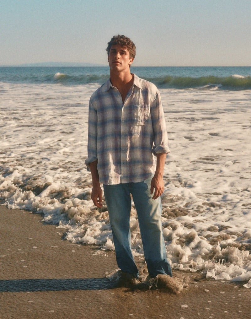 Kai stands in the shore break wearing a blue ombre plaid linen shirt with the sleeves rolled and light-wash jeans