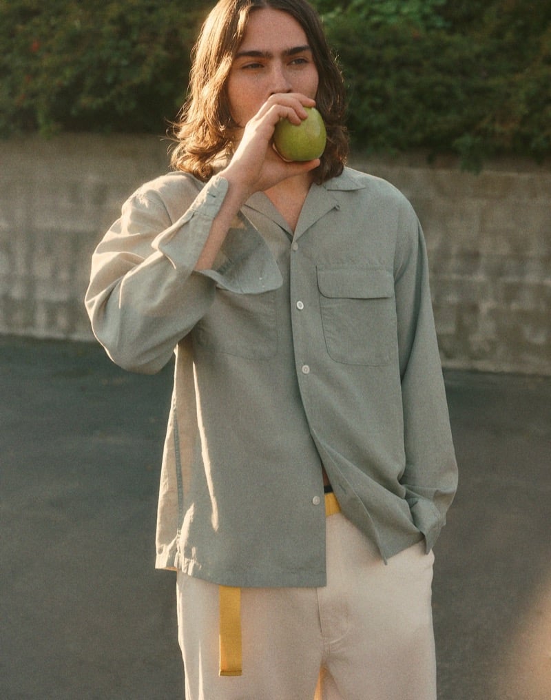 Sava bites into a green apple wearing a sage-green camp shirt with double chest pockets and cream trousers with a yellow belt