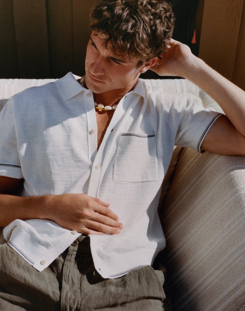Kai reclines on a striped sofa in a white camp shirt with dark navy tipping on the collar and cuffs and khaki linen shorts with a shell necklace