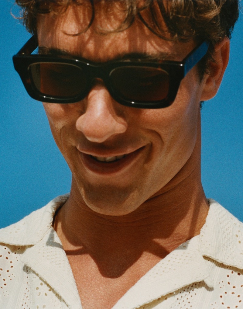 Kai smiles in a close-up wearing black rectangular sunglasses and an eyelet-embroidered cream camp shirt against a blue sky