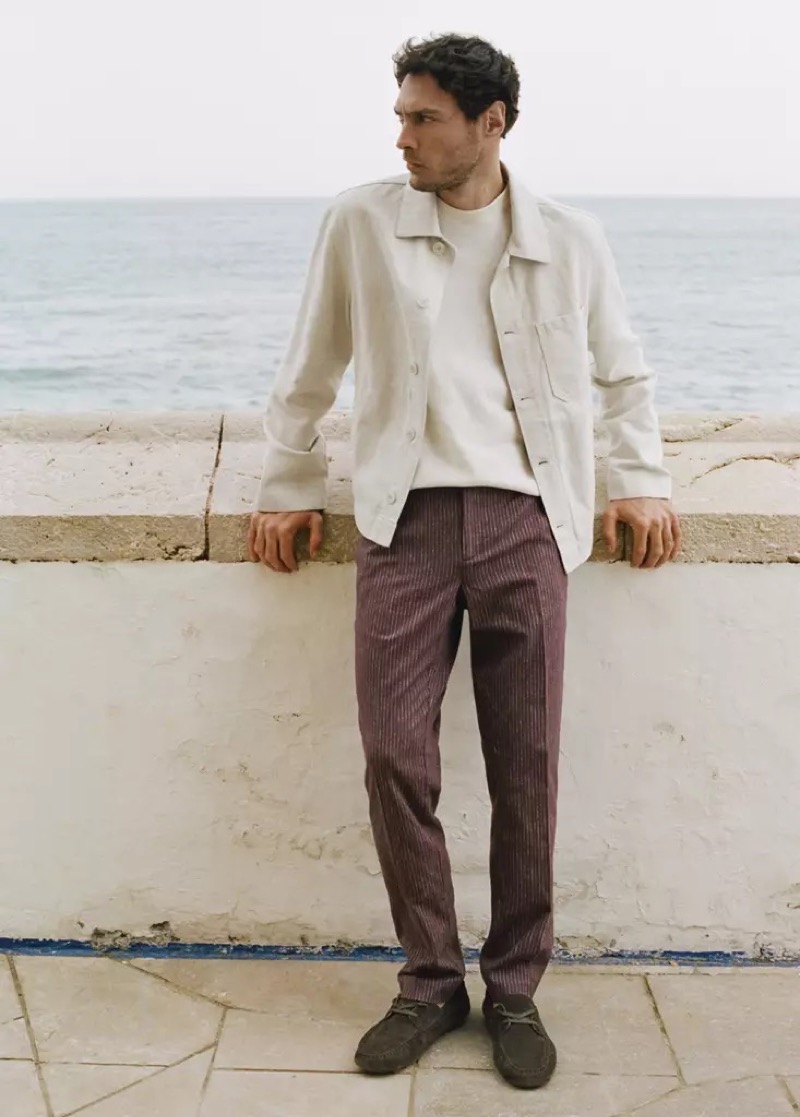 Pierre-Benoit wears a cream jacket over a tee with brown striped trousers at a sea wall