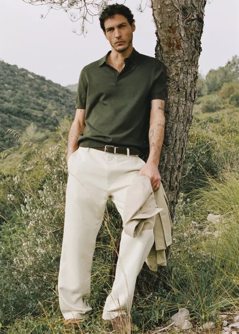 Pierre-Benoit wears a dark sage knit polo with cream trousers holding a jacket