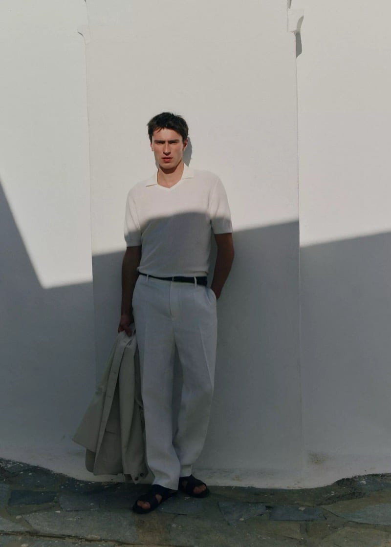 Clément stands against a sunlit white wall in a cream knit polo, wide-leg white linen trousers, and black sandals, holding a beige jacket, for Mango Selection's spring-summer 2026 collection
