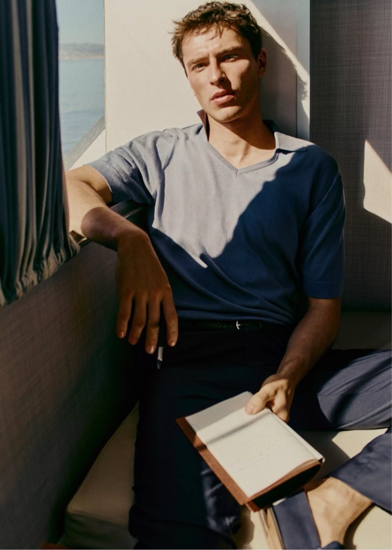 Clément reclines on a sofa by a sea view in a blue knit polo and dark trousers, holding an open book, for Mango Selection's spring-summer 2026 collection