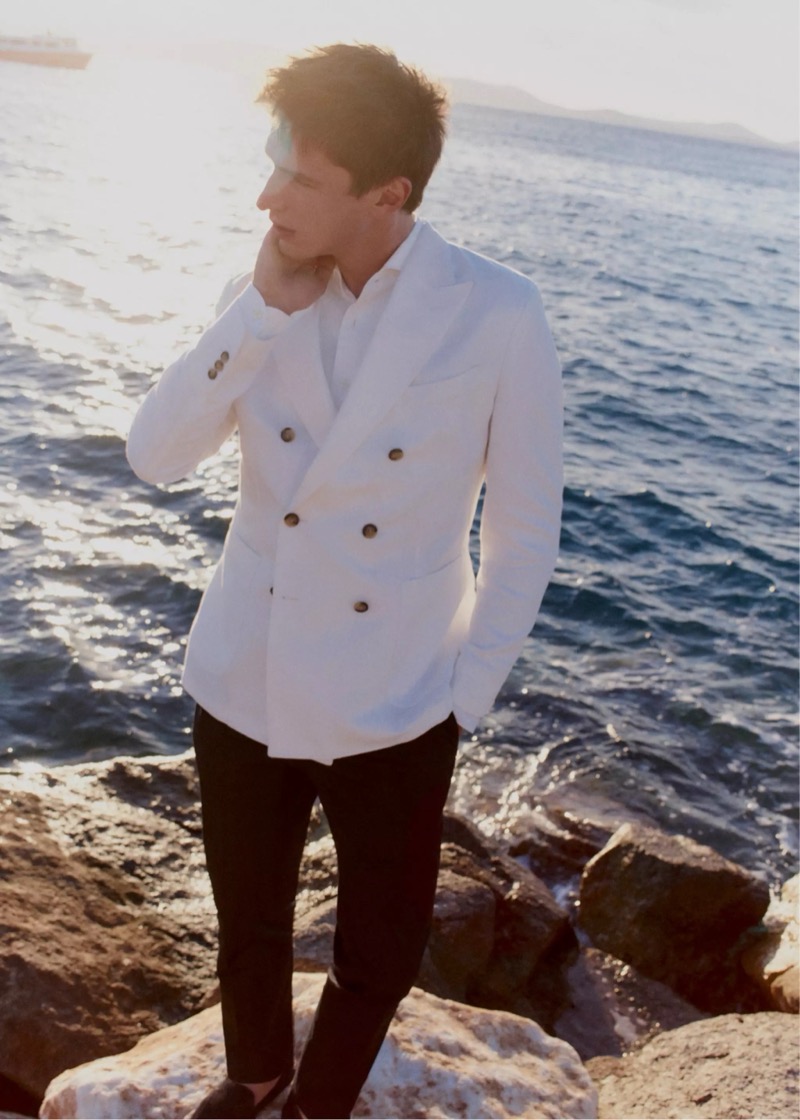 Clément stands on coastal rocks at sunset in a white double-breasted linen jacket over a white shirt and dark trousers for Mango Selection's spring-summer 2026 collection
