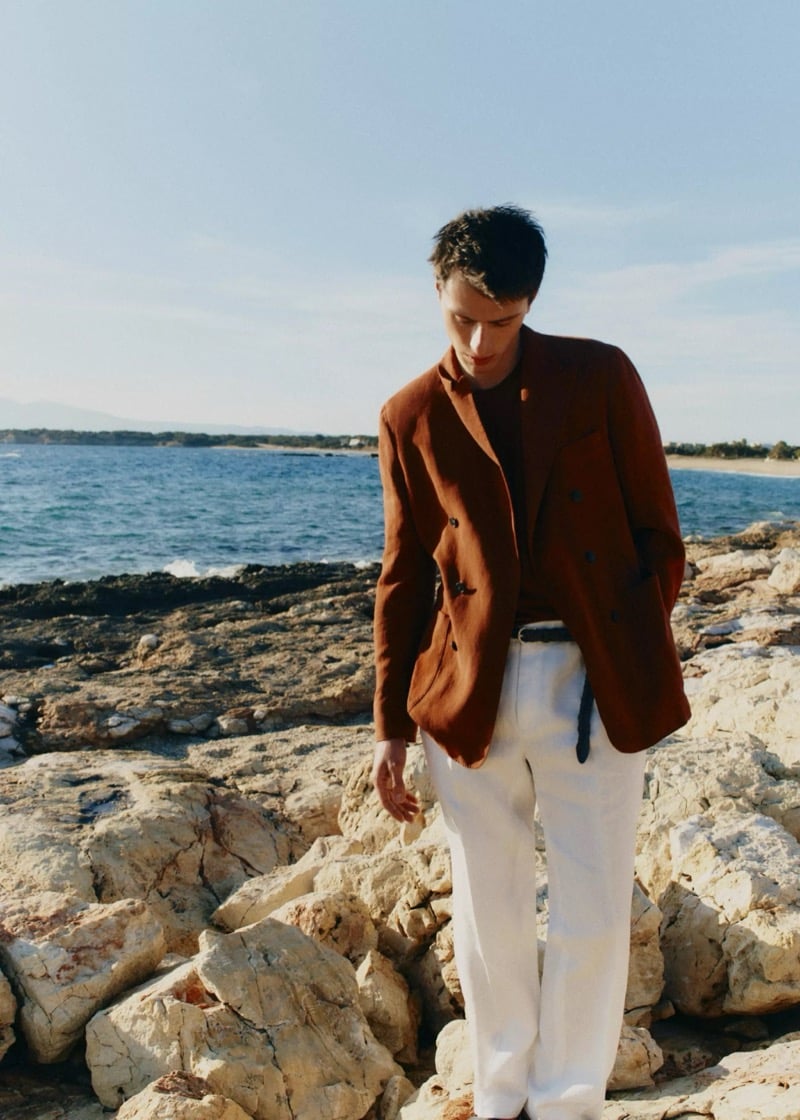 Clément stands on a rocky coast in a rust double-breasted linen blazer over a knit polo and wide-leg white linen trousers for Mango Selection's spring-summer 2026 collection