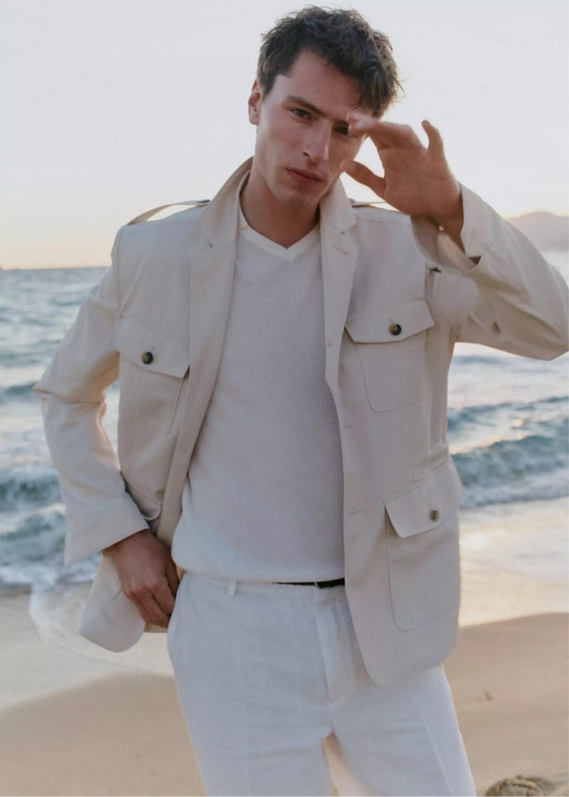 Clément stands on a beach at sunset in a cream cotton field jacket layered over a white V-neck knit and white linen trousers for Mango Selection's spring-summer 2026 campaign