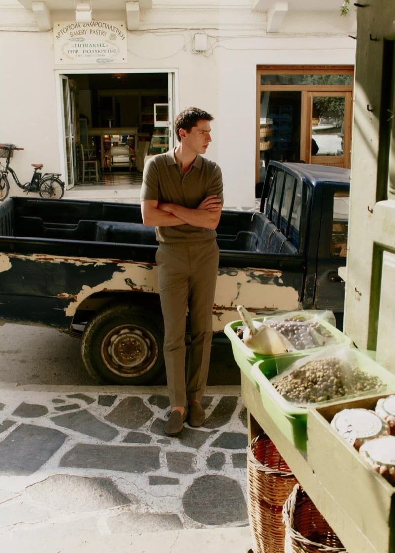 Clément stands with arms crossed by a vintage pickup on a Greek street in an olive knit polo, olive trousers, and suede loafers for Mango Selection's spring-summer 2026 collection