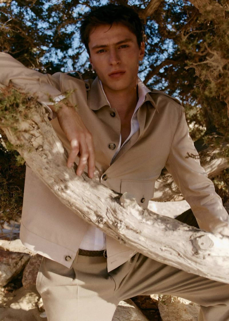 Clément leans through dry coastal trees in a beige cotton field jacket over a white shirt with cream trousers for Mango Selection's spring-summer 2026 collection