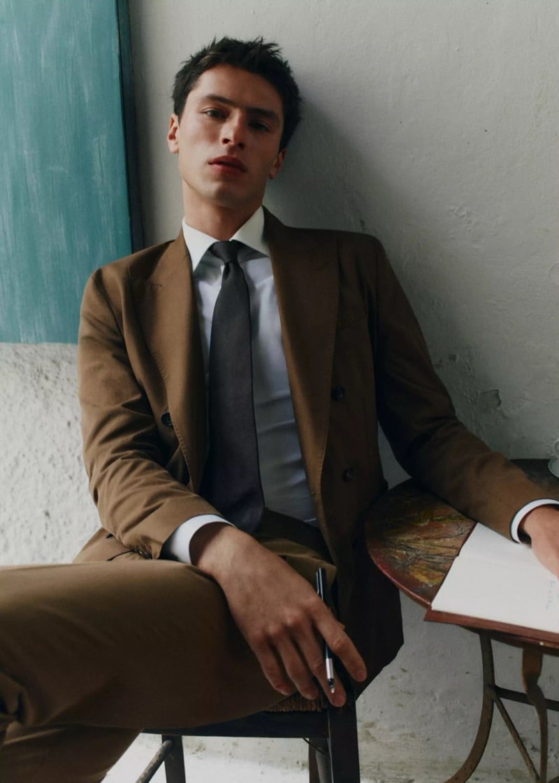 Clément sits in a brown suit with a white shirt and gray tie at a small painted table for Mango Selection's spring-summer 2026 campaign