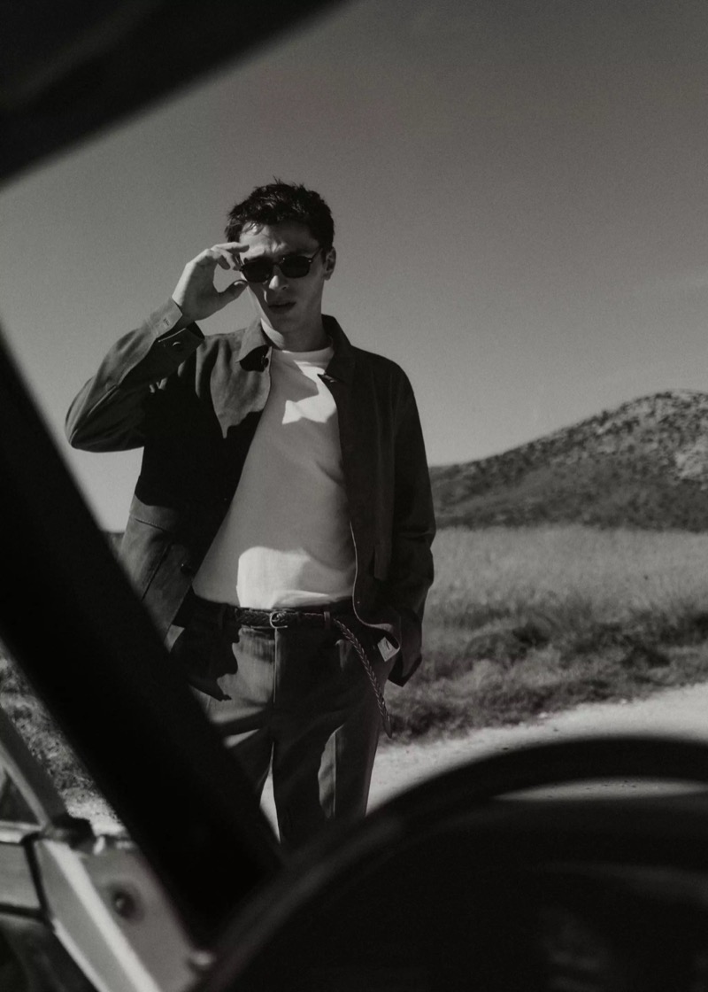 Black and white image of Clément in sunglasses standing beside a vintage car on a dirt road in a dark jacket, white tee, and dark trousers for Mango Selection's spring-summer 2026 collection